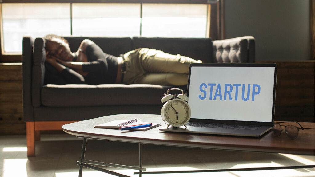 Image of person napping on couch, with alarm clock and laptop in the foreground